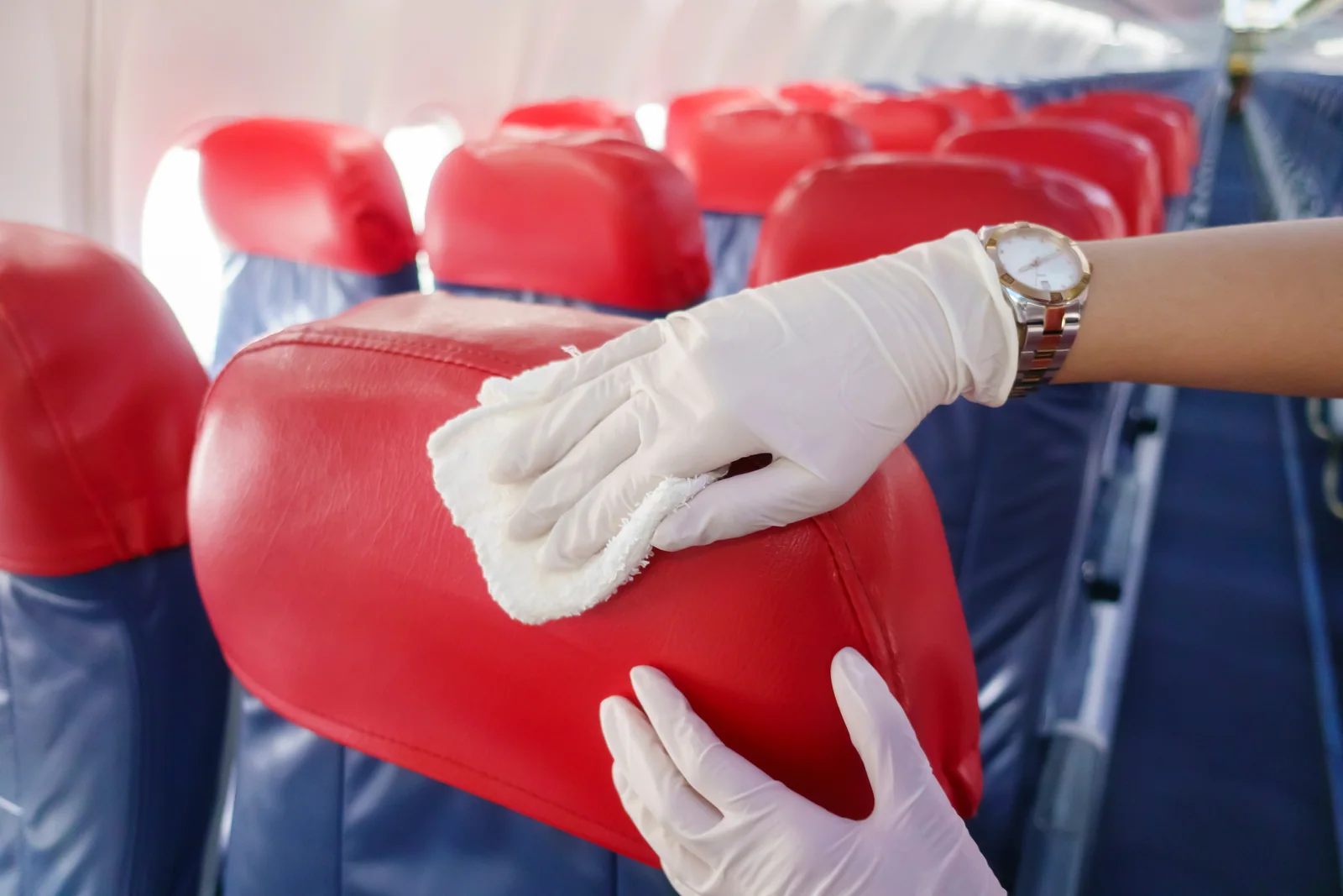 Gloved hands cleaning a red aircraft seat with a cloth.